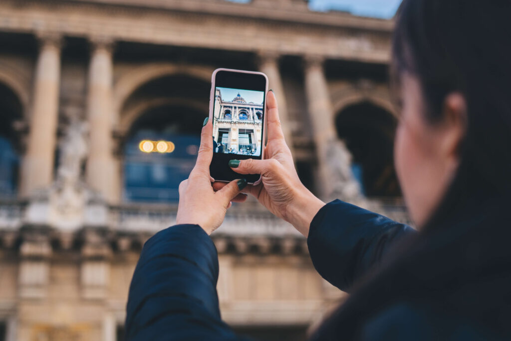 Dłonie trzymają smartfon, na którego ekranie widać zabytkowy budynek z kolumnami w tle.